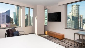 A modern hotel room with large windows shows a cityscape view, featuring a TV, chair, and minimalist decor.