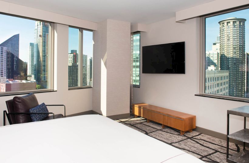 A modern hotel room with large windows shows a cityscape view, featuring a TV, chair, and minimalist decor.