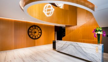 The image shows a modern lobby with a marble desk, wooden walls, a large clock, spiral design, and decorative lighting.