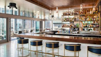A modern bar with high stools, a polished countertop, and a wide selection of liquor bottles on shelves against a city view backdrop.