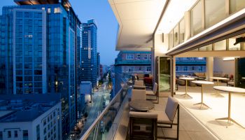 A modern cityscape at dusk with a lit rooftop terrace, featuring seating, tables, and a view of tall buildings and a street.