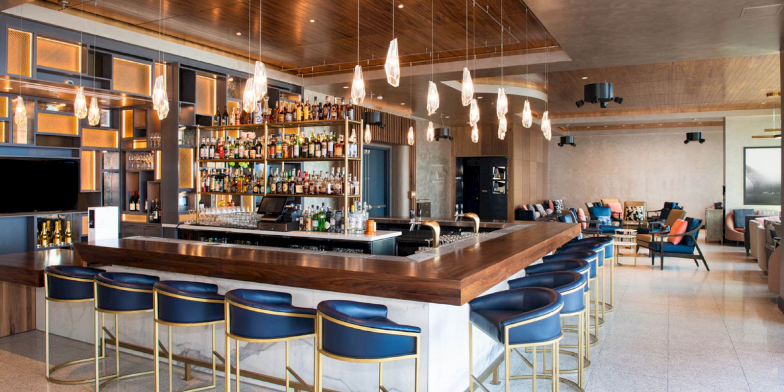 A modern bar with a wooden countertop, blue stools, hanging lights, and a well-stocked back shelf in a spacious lounge area.