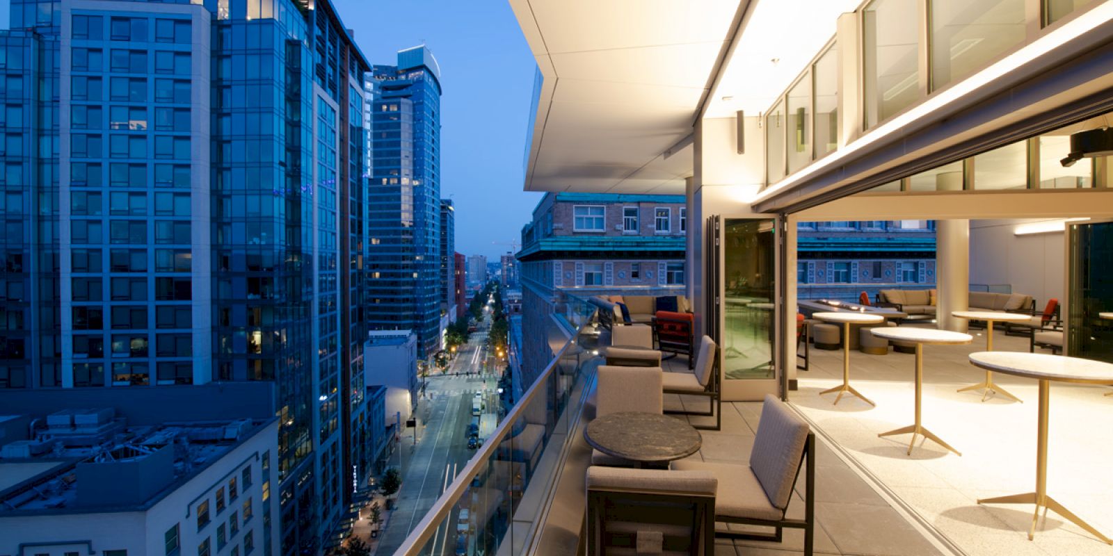 A modern cityscape with tall buildings, seen from a balcony with seating and tables, illuminated by evening lights.