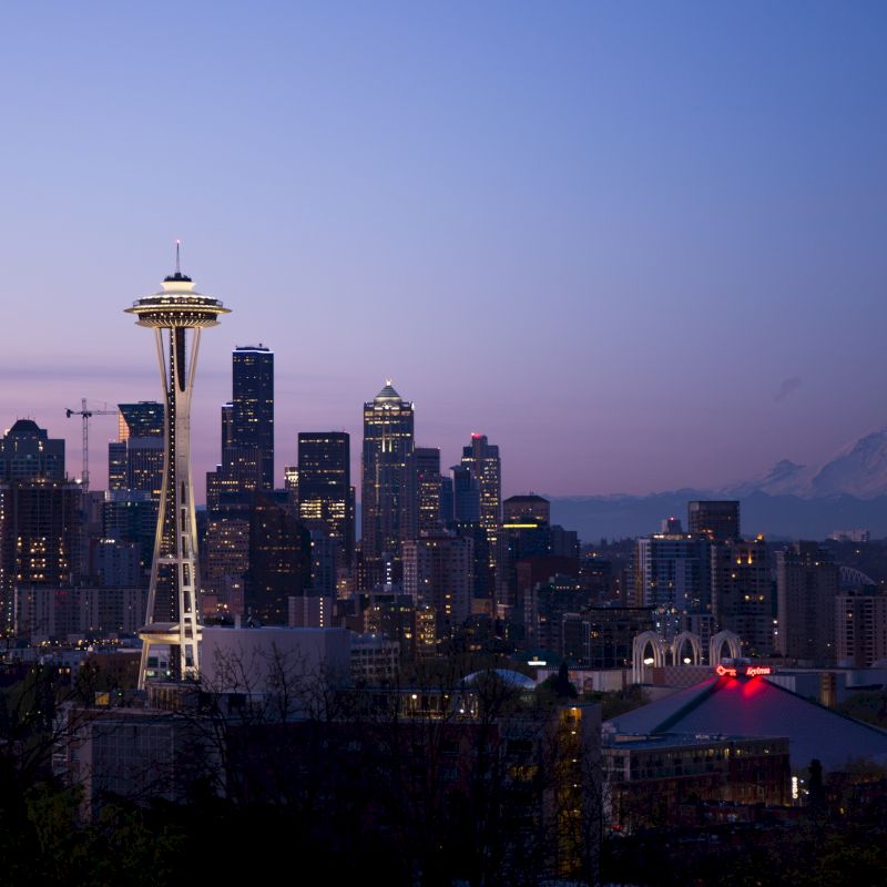 Seattle skyline at dusk with the Space Needle prominent, city buildings, and Mount Rainier in the background under a clear sky.