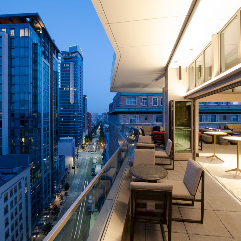 A modern rooftop terrace with seating overlooks a cityscape, featuring tall glass buildings under an evening sky.