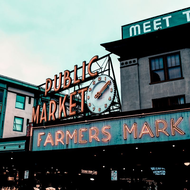 The image shows a neon sign reading "Public Market" and "Farmers Market" with a clock, part of an iconic marketplace setting.