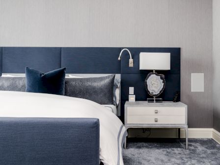 A modern bedroom with a navy bed, white bedding, and a bedside table with a lamp and decorative stone, set against a light gray wall.