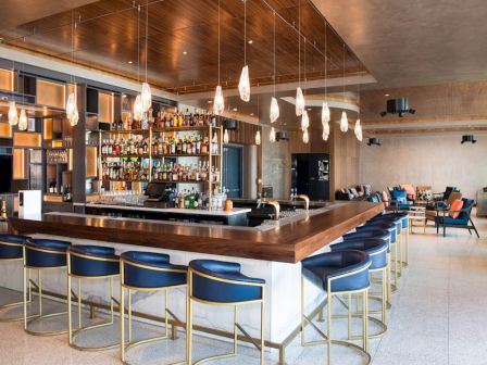 A modern bar with hanging lights, blue barstools, and a well-stocked counter. Comfortable seating is in the background.