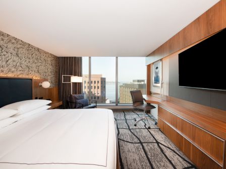 A modern hotel room with a large bed, wall-mounted TV, desk, and floor-to-ceiling window with a city view.