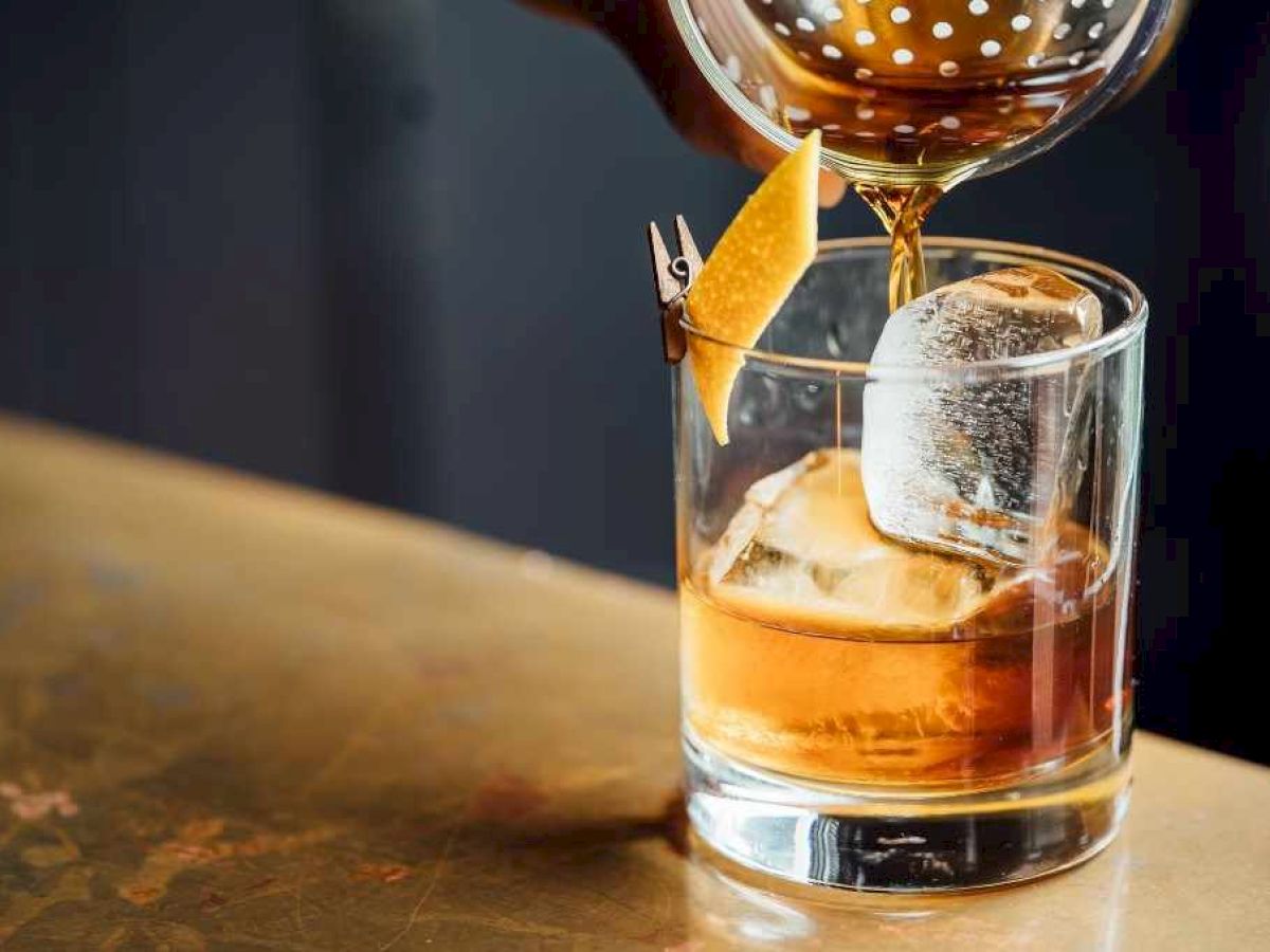 A cocktail is being poured into a glass with ice and a lemon peel garnish on a bar top.