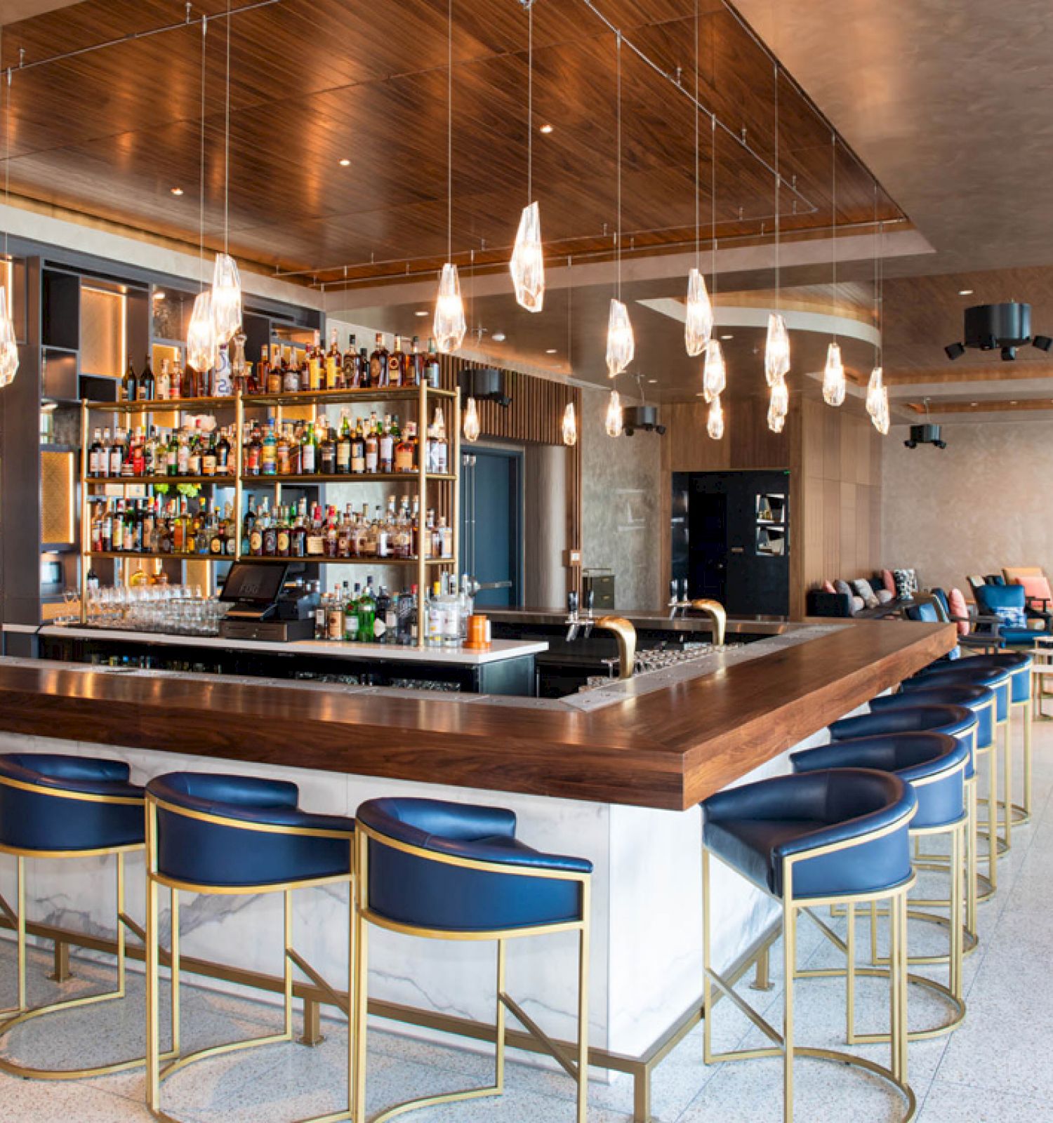 A modern bar with a wooden counter, blue stools, hanging lights, and a fully stocked back shelf, situated in a spacious lounge area.
