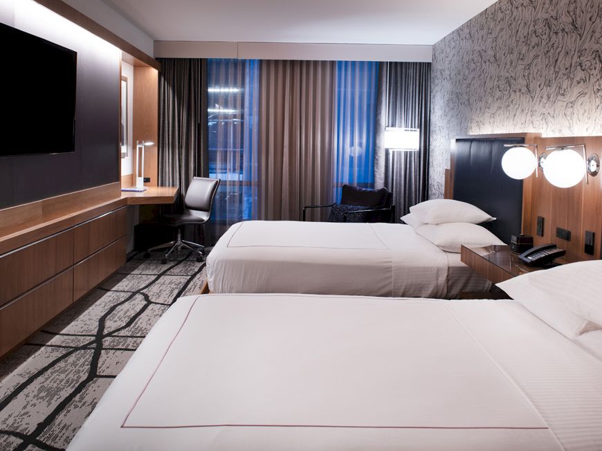 A modern hotel room with two beds, a flat-screen TV, desk, and lamp, featuring patterned wall and carpet, and large window with curtains.