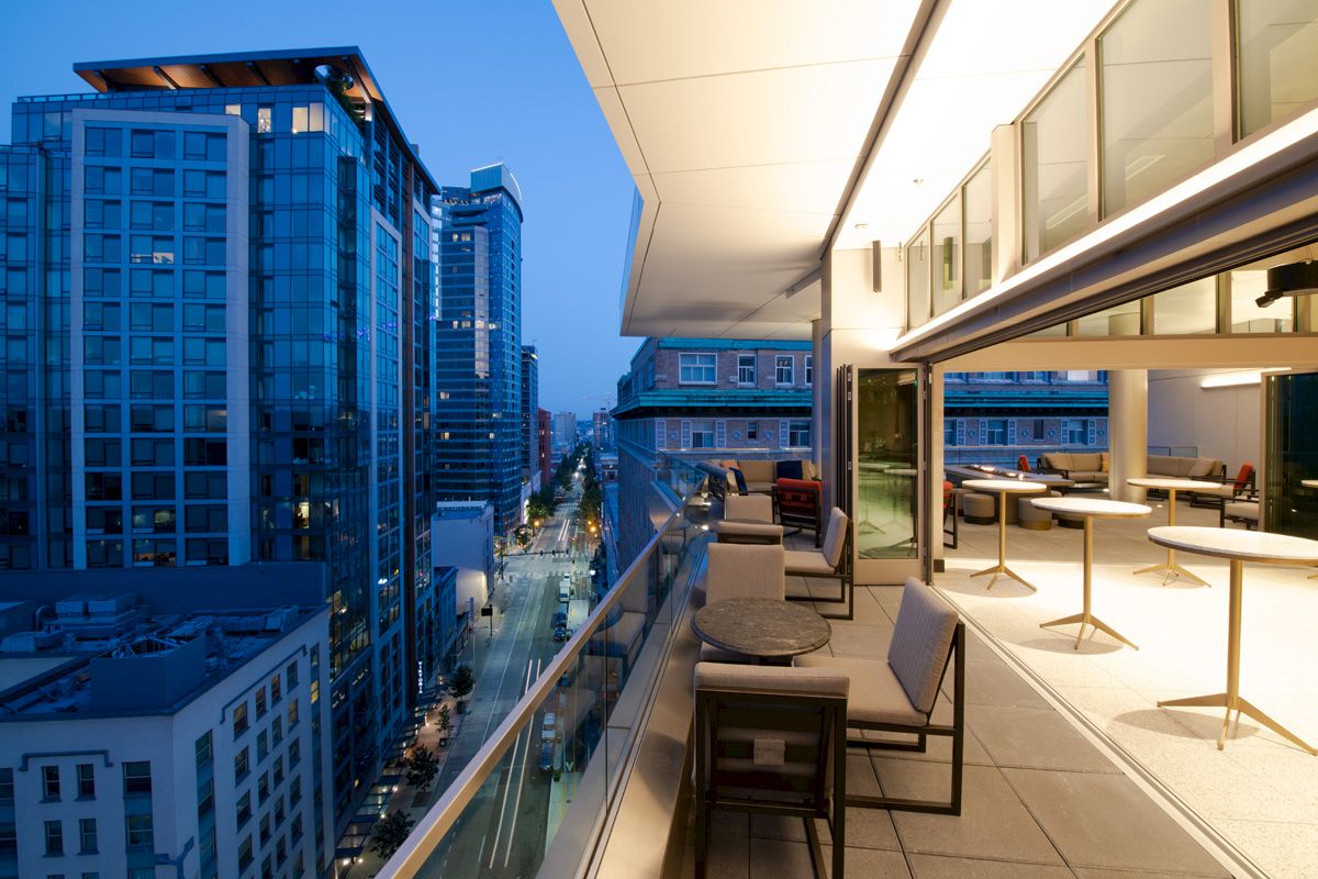 The image shows a modern cityscape with tall buildings and a sleek balcony featuring seating and tables, overlooking an urban street.