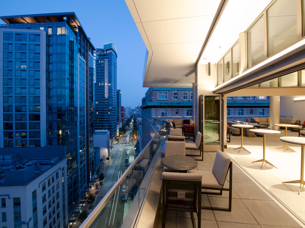 The image shows a modern cityscape with tall buildings and a sleek balcony featuring seating and tables, overlooking an urban street.