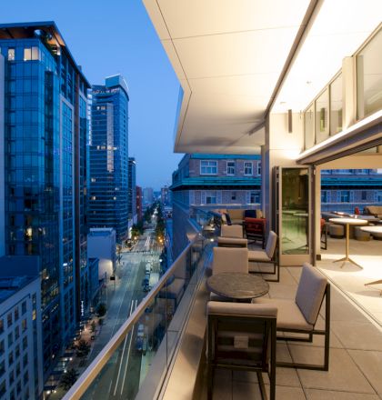 A modern rooftop patio overlooks a city street, surrounded by tall buildings at dusk, with outdoor seating and tables.