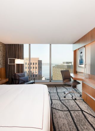 A modern hotel room features a large bed, desk, TV, chair, and a window with an urban view.
