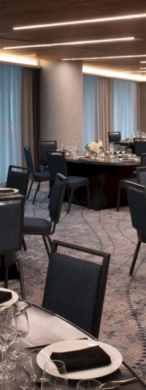 The image shows an elegantly set dining room with round tables, glassware, and floral centerpieces, under soft lighting and large windows.