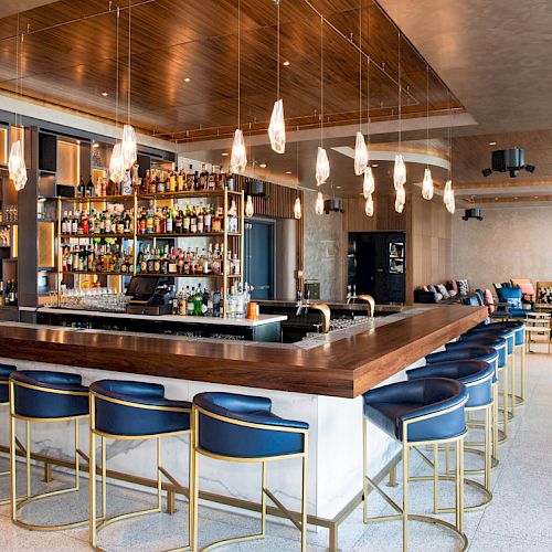 A stylish bar with a wooden counter, blue stools, hanging lights, and a well-stocked liquor shelf, set in a modern lounge area.