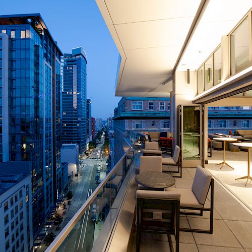 A rooftop terrace with seating overlooks a cityscape at dusk, with modern high-rise buildings lining the street below.
