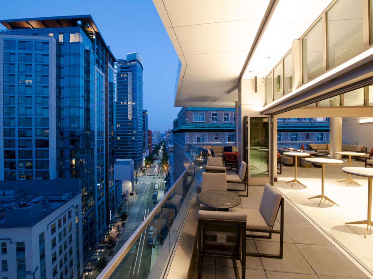A rooftop terrace with seating overlooks a city street with modern buildings at twilight.