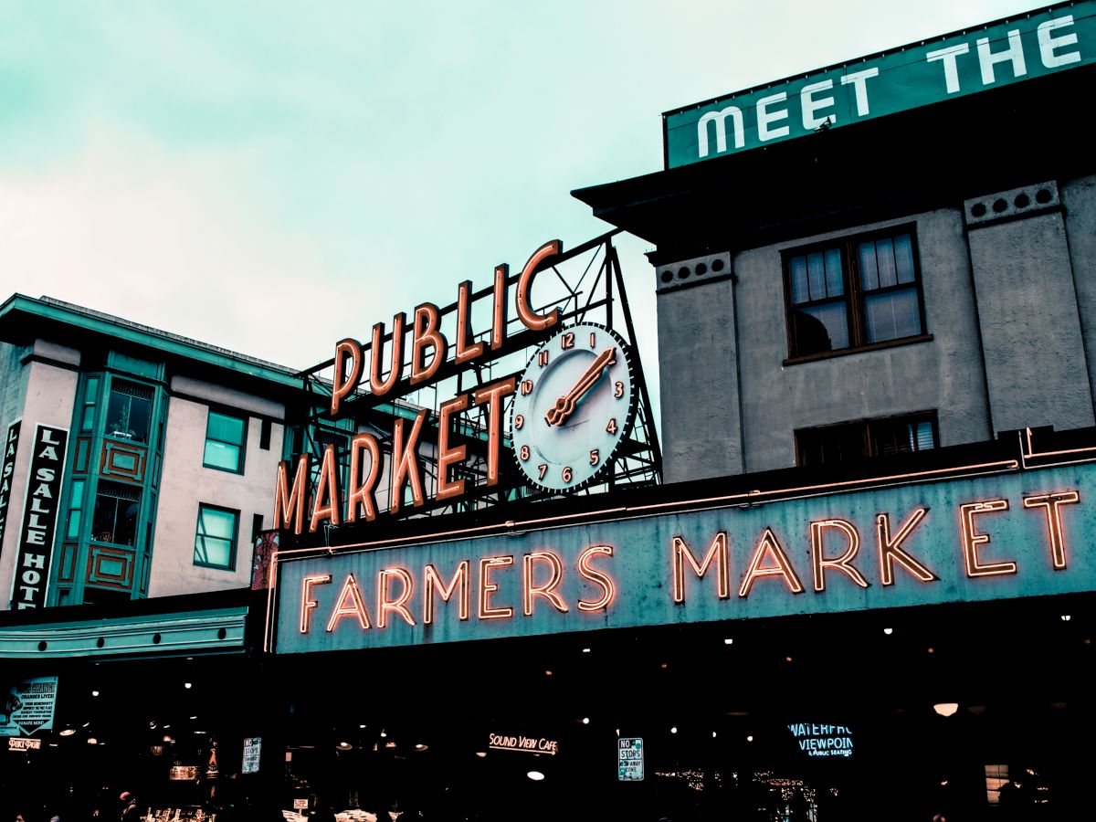 The image shows a neon sign for a public farmers market, featuring a clock, against a background of buildings under a cloudy sky.