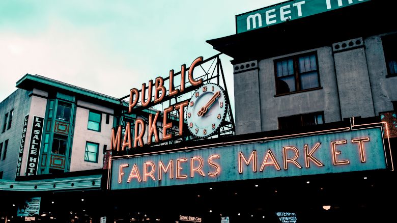 The image shows a neon sign for a public farmers market, featuring a clock, against a background of buildings under a cloudy sky.