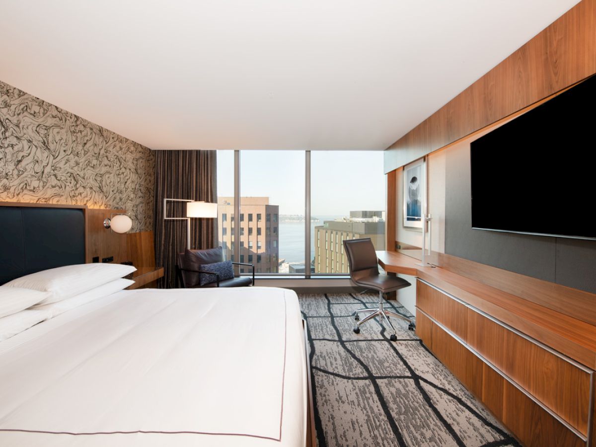 A modern hotel room features a double bed, large TV, desk, chair, and window with a city view, creating a sleek and cozy atmosphere.