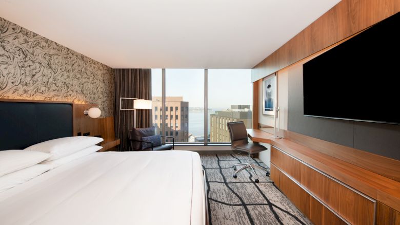 A modern hotel room features a double bed, large TV, desk, chair, and window with a city view, creating a sleek and cozy atmosphere.