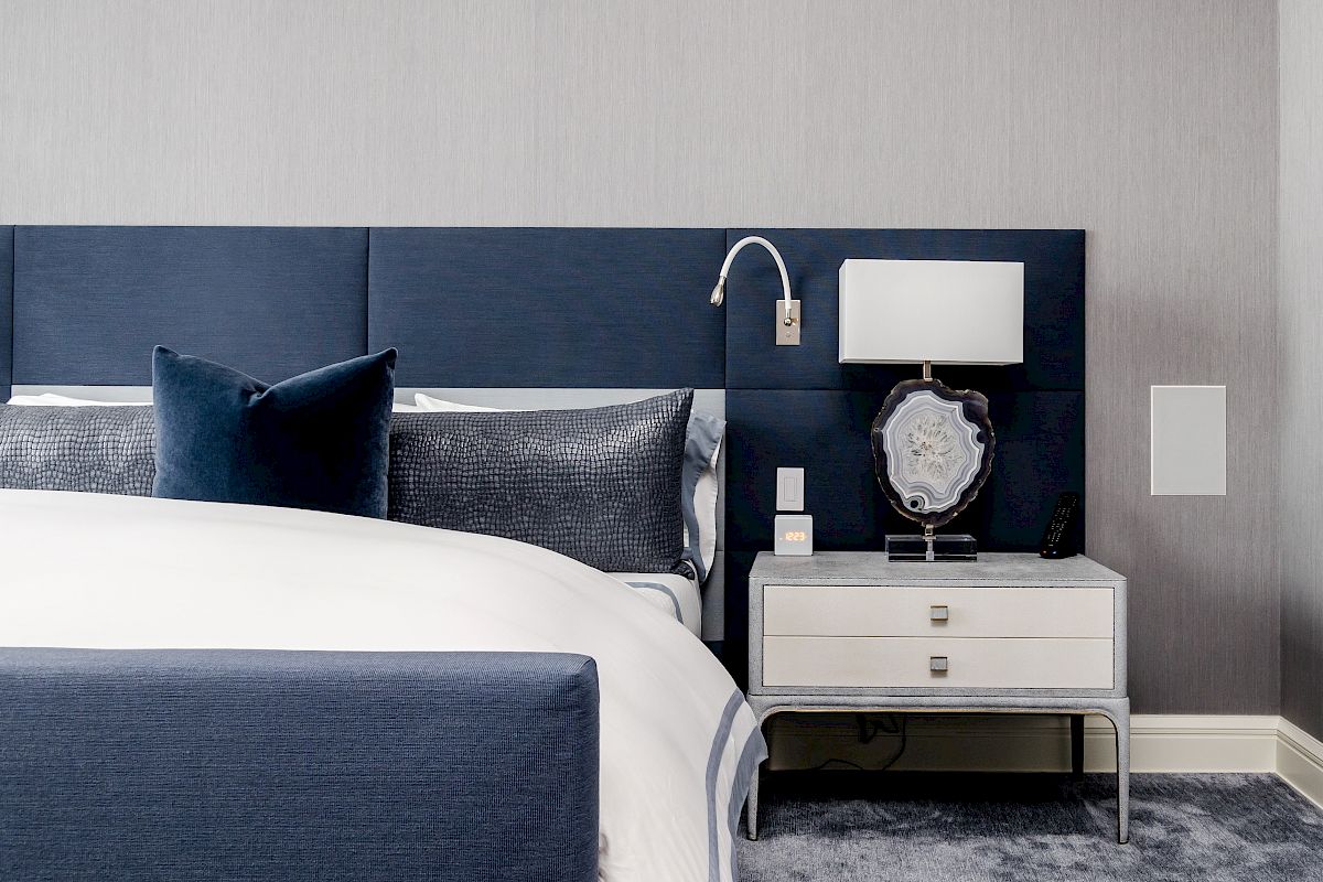 A modern bedroom with a navy upholstered headboard, bedside table, lamp, and white and gray bedding, featuring a clean and elegant design.