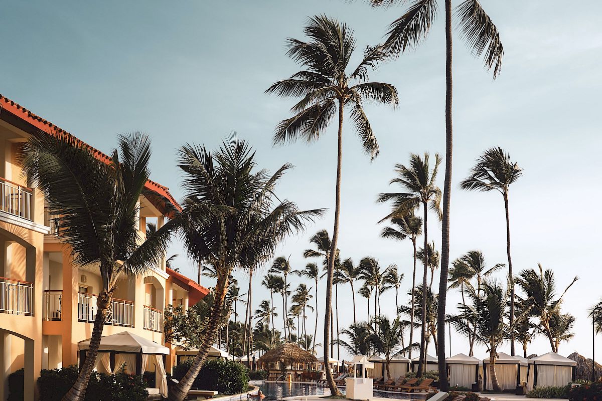 A luxurious poolside scene with sun loungers, tall palm trees, and a resort building under a clear sky.