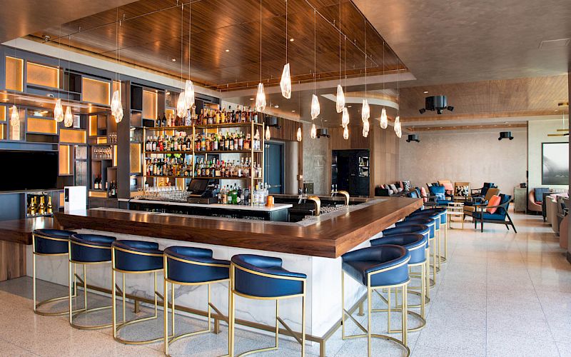 A modern bar with a fully stocked back wall, pendant lights, and blue bar stools around a wooden counter in a spacious lounge area.