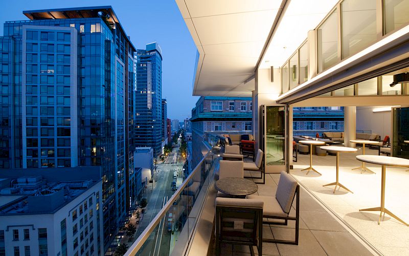 A modern cityscape at dusk with high-rise buildings and a stylish balcony featuring seating and tables.